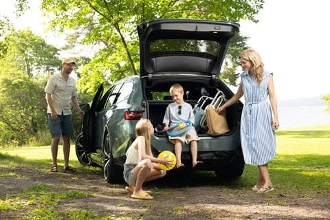 Elbil-SUV i sommerhage. Kone og mann står på hver sin side av bilen. Et gutt sitter i bagasjerommet, og en jente sitter på bakken og ser opp på gutten.