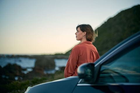 Mulher encostada ao carro a olhar para o pôr do sol