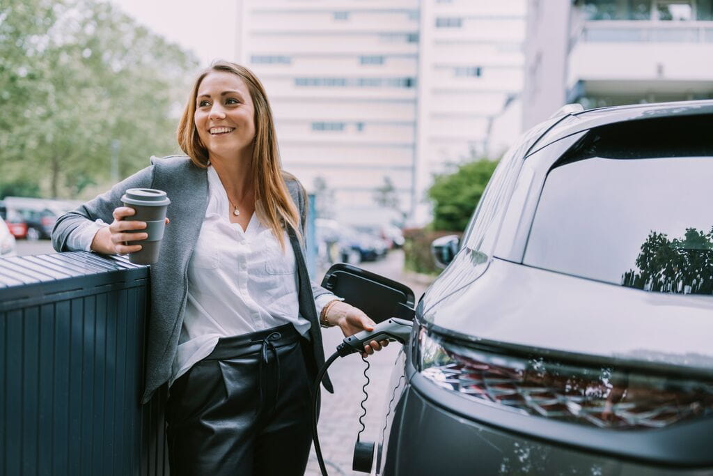 Happy woman with disposable coffee cup charging car at station